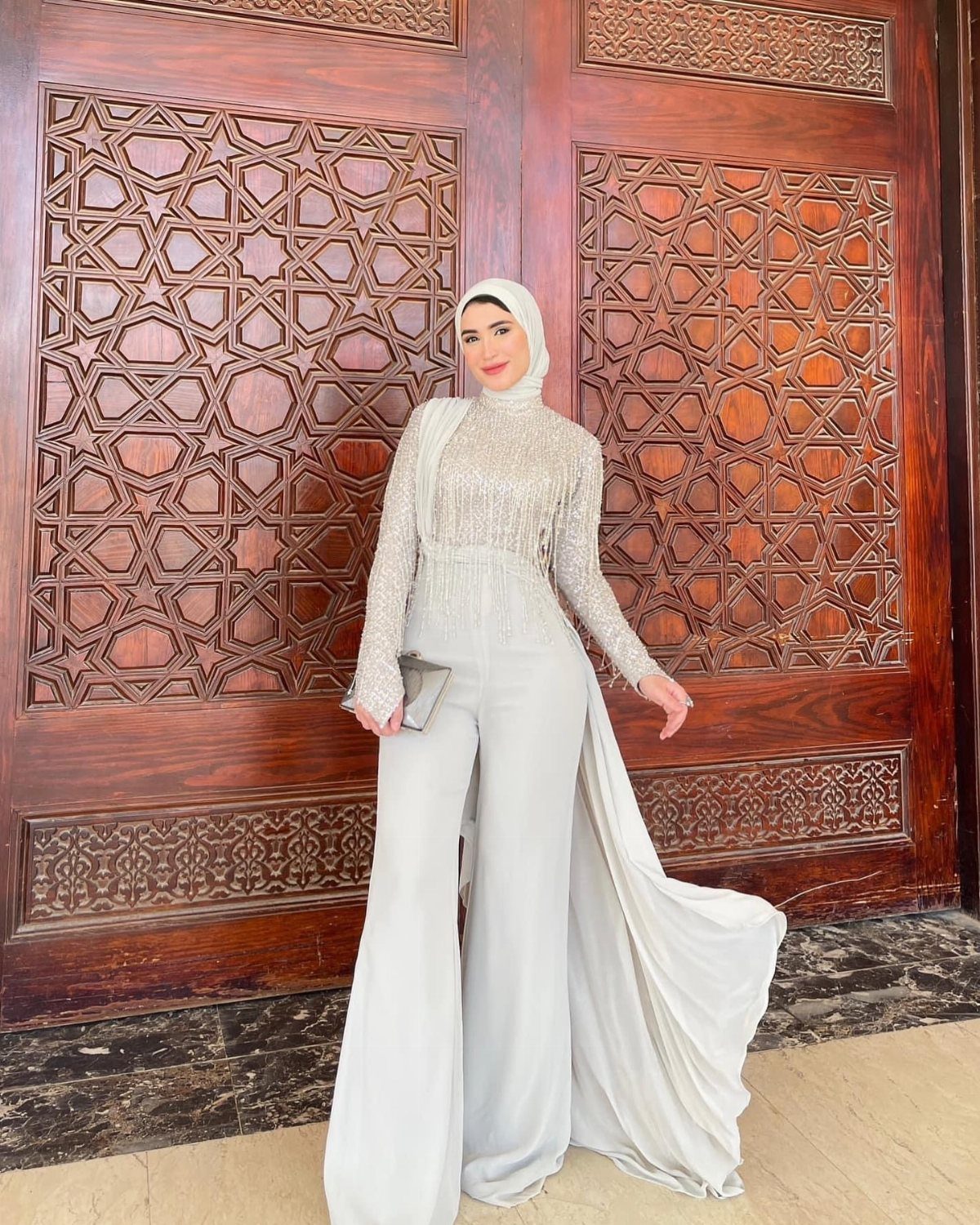 Woman in a shimmering grey wide-leg jumpsuit and hijab standing by carved doors.