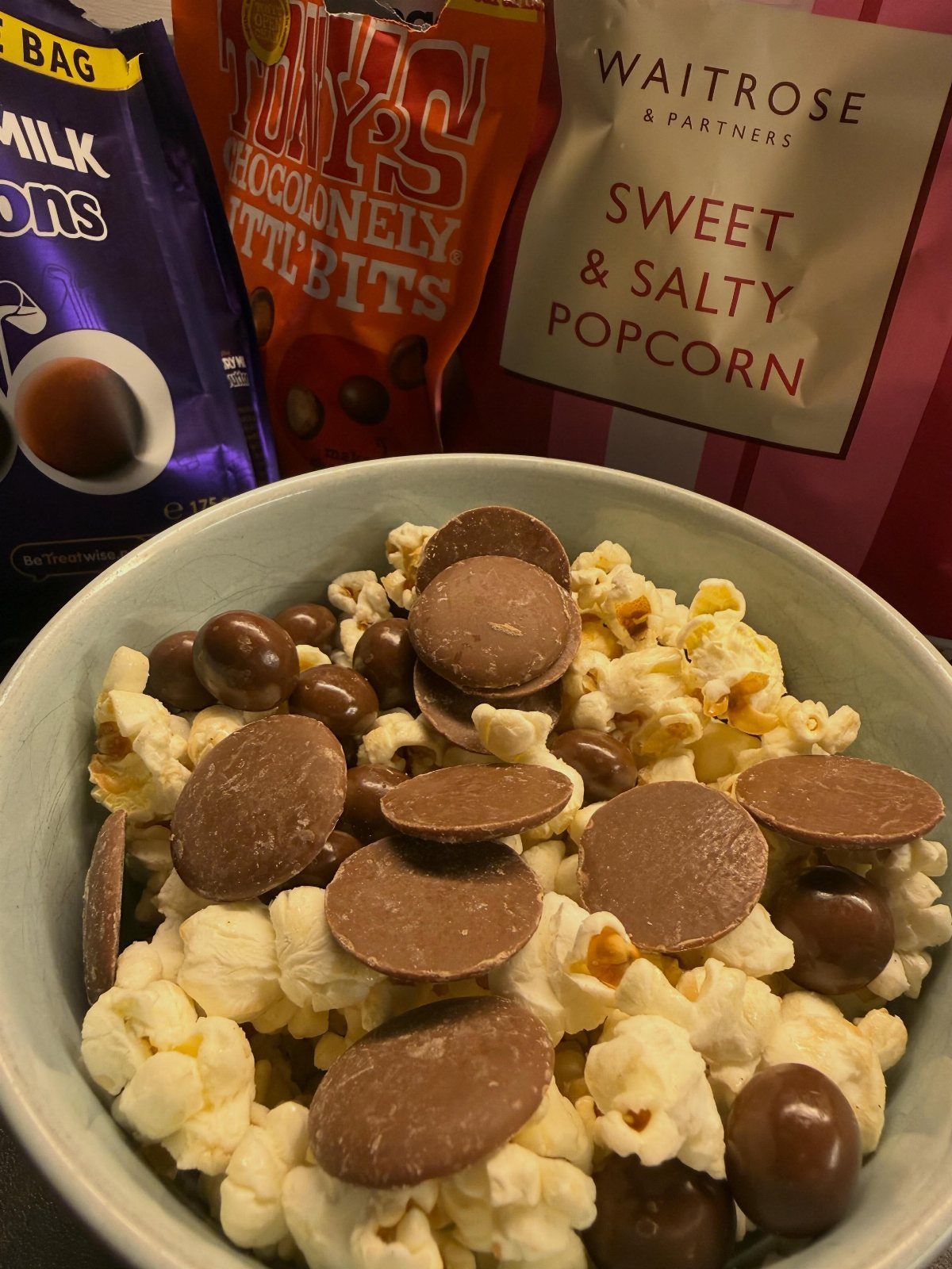 Bowl of popcorn mixed with chocolate discs and balls next to snack bags.