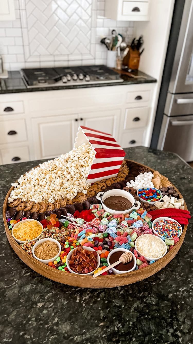 Snack platter with spilling popcorn bucket and assorted candies on a kitchen counter.