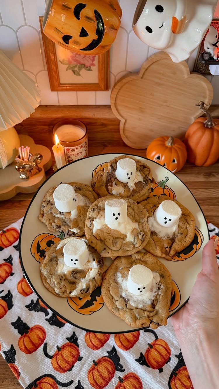 Ghost-themed chocolate chip cookies on a plate surrounded by Halloween pumpkins and decor.