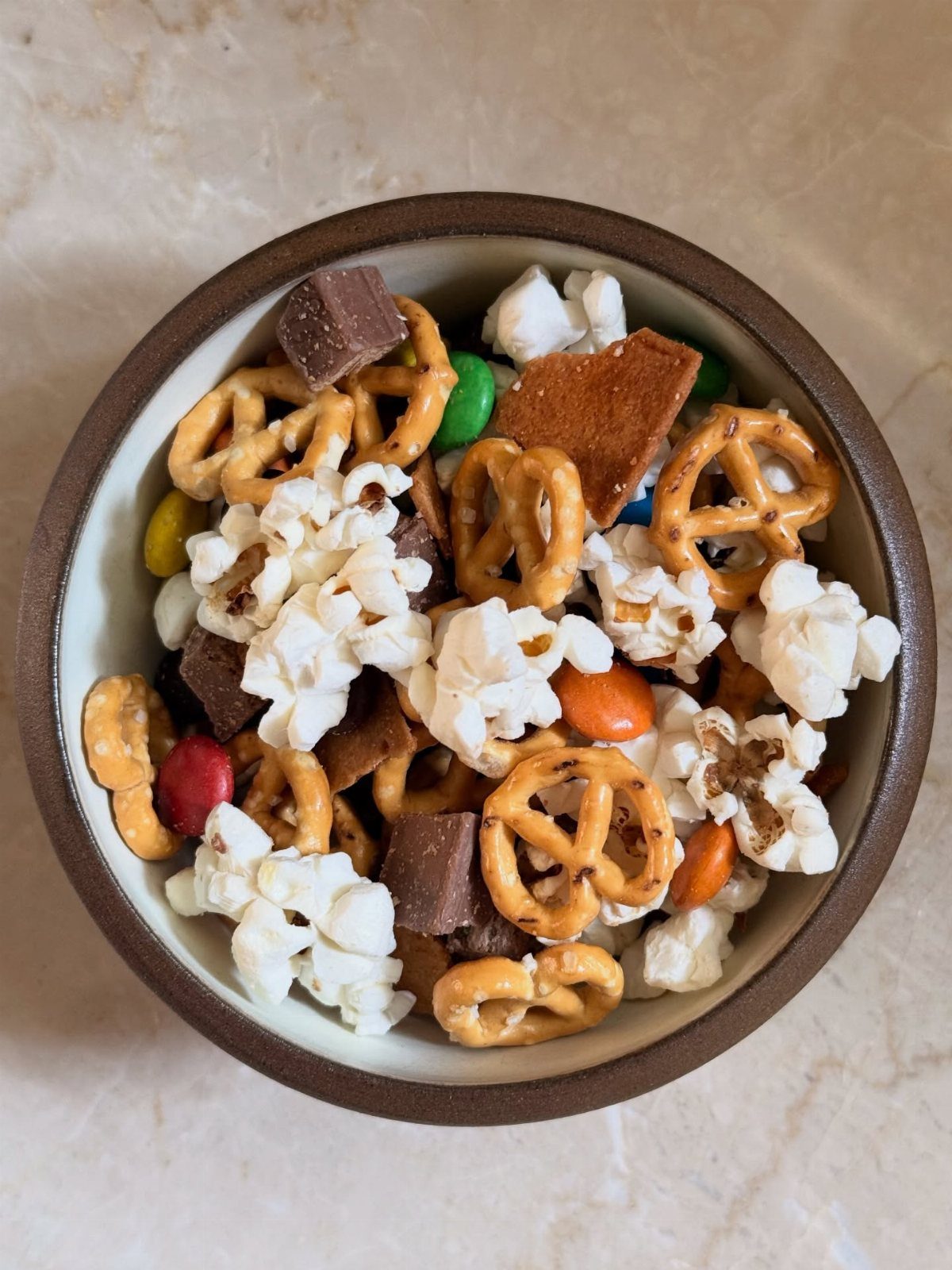 Snack mix with popcorn, pretzels, and chocolate in a brown ceramic bowl.