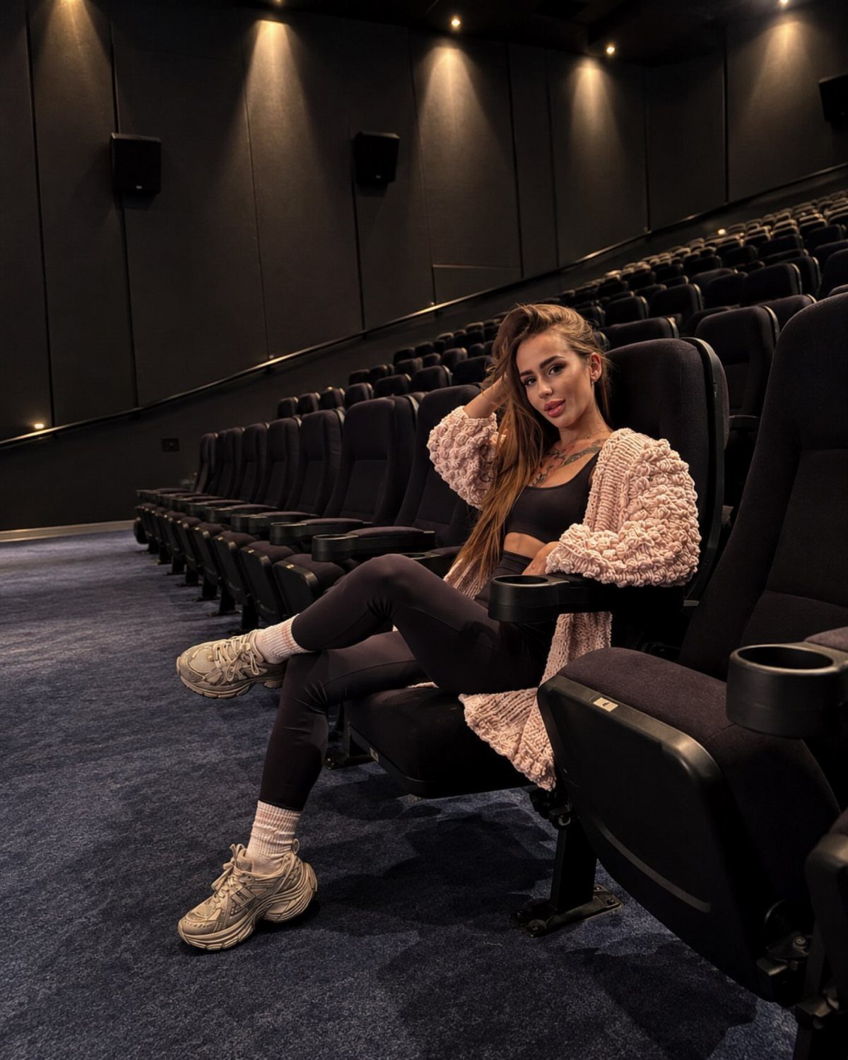 Woman in a pink knitted cardigan and black leggings sitting in cinema seats.