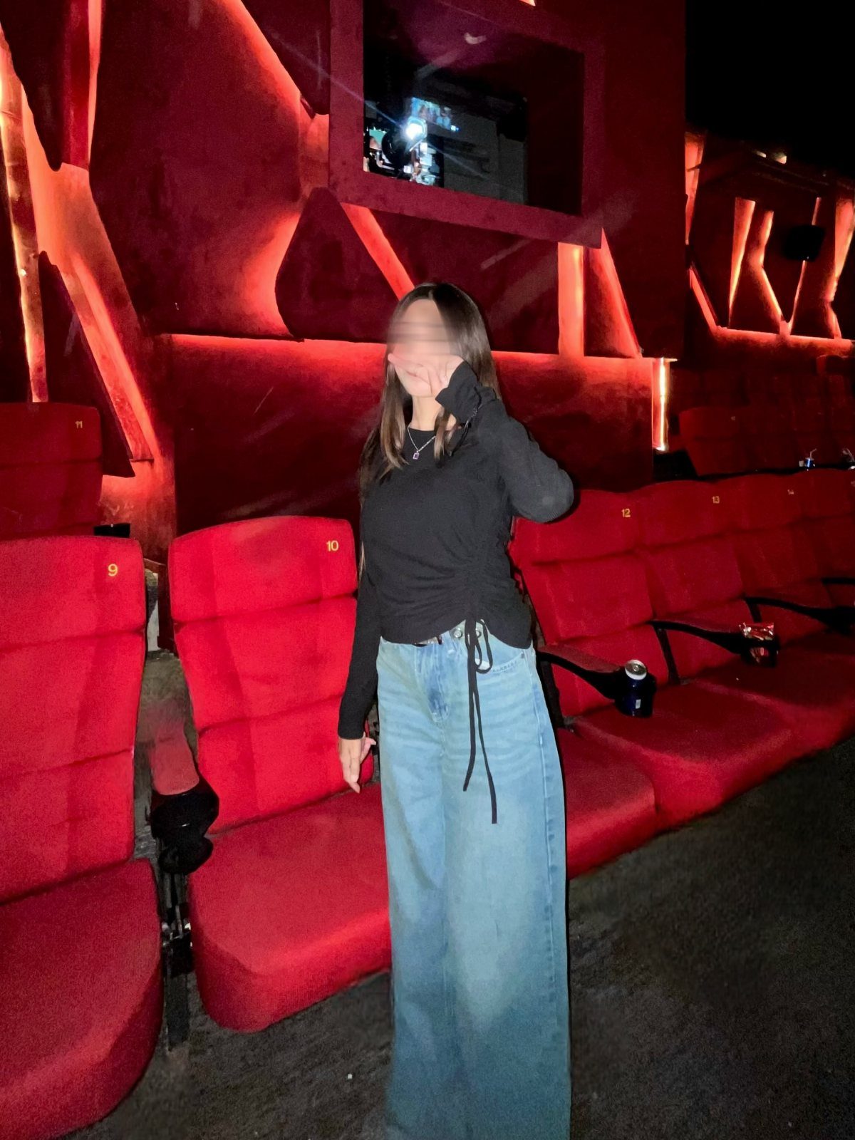 Woman in a black top and wide-leg denim jeans standing in red cinema seats.