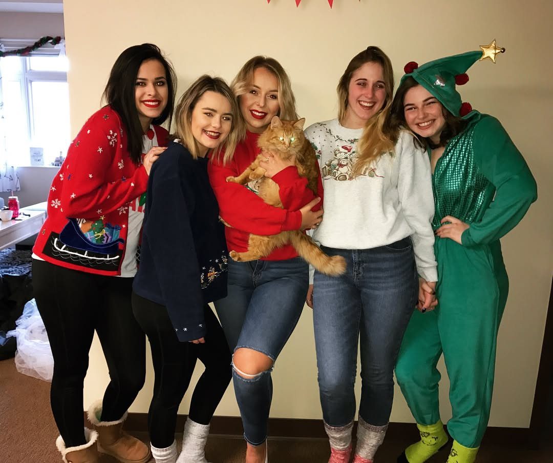 Five women in cozy sweaters and a green elf onesie posing together indoors.