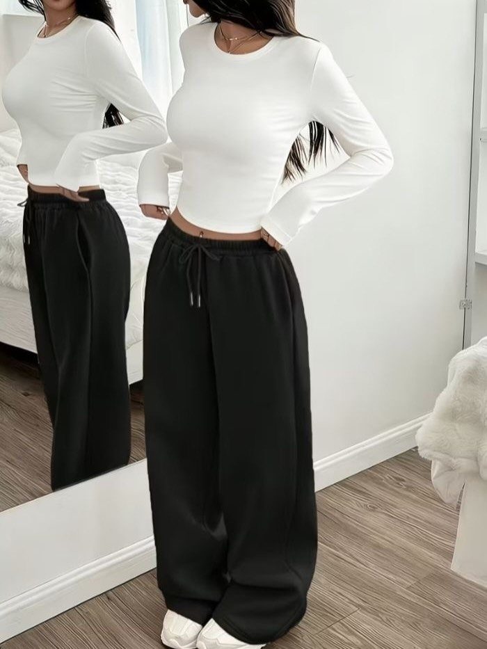 Woman wearing a white long-sleeved crop top and baggy black joggers standing by mirror.