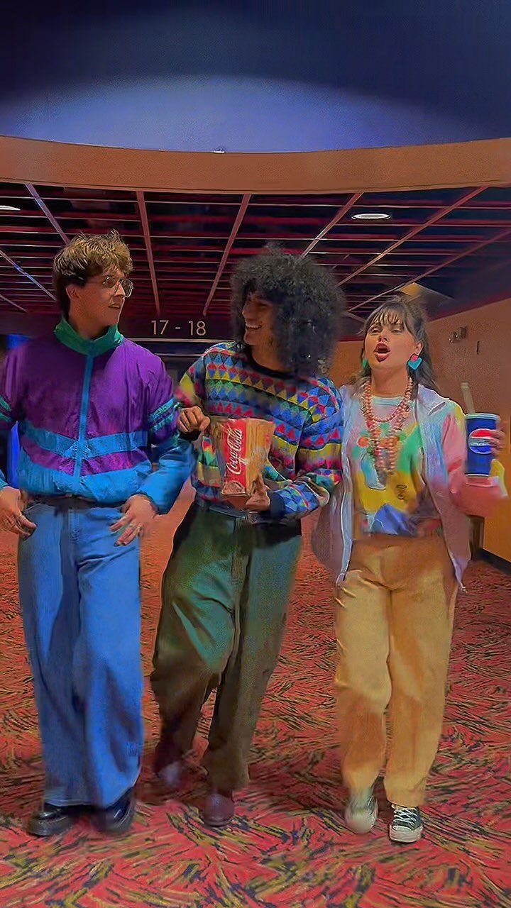 Three people in colorful 80s outfits standing in a movie theater lobby.