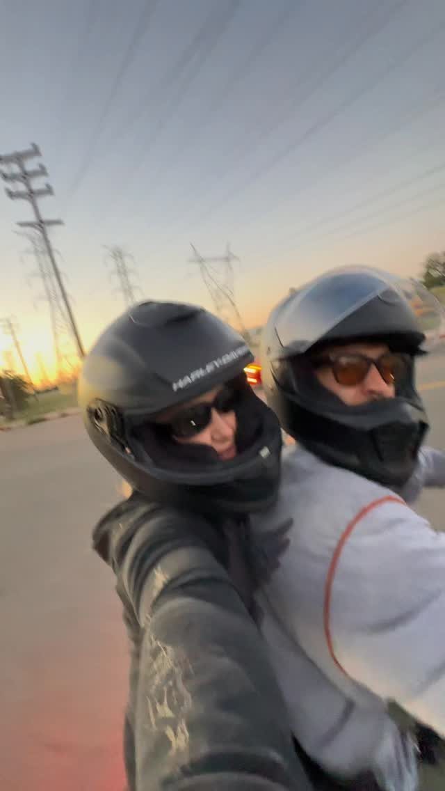 Couple wearing motorcycle helmets taking a selfie while riding a motorcycle at sunset.