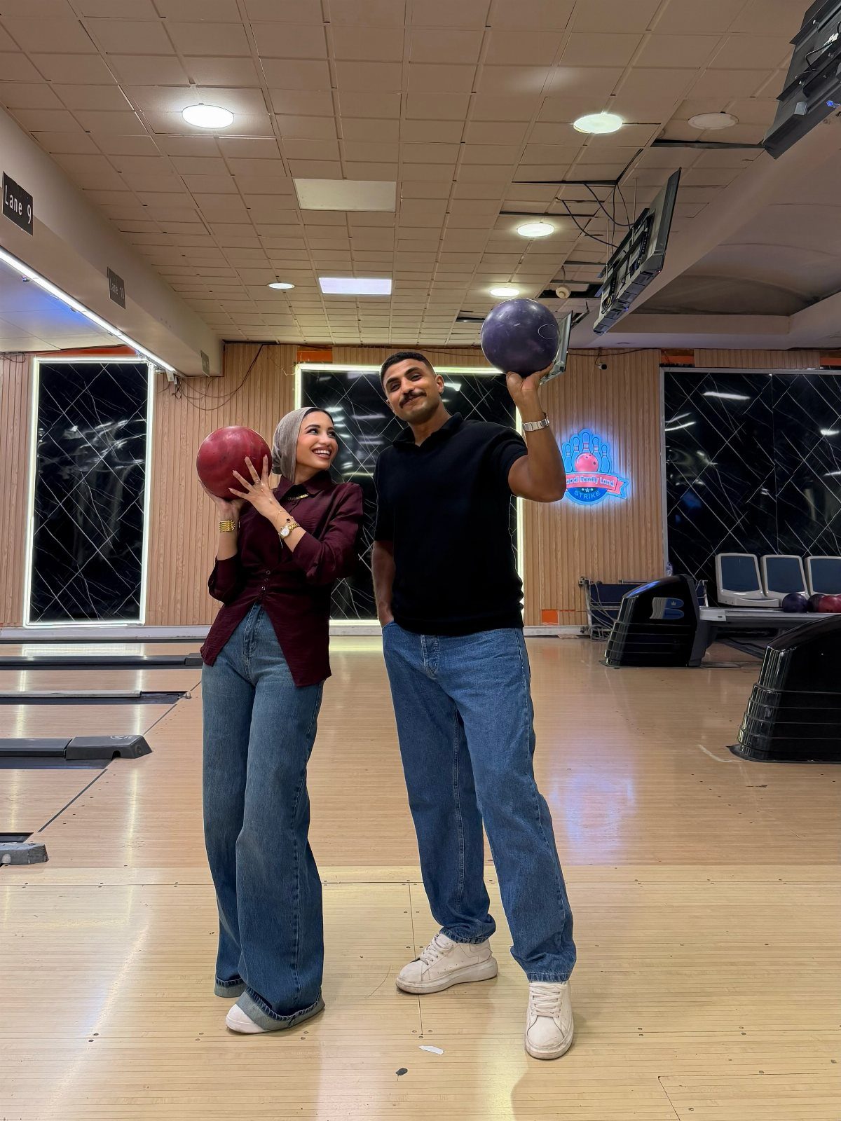 A couple posing with bowling balls in a brightly lit bowling alley.