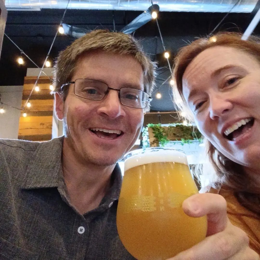 Smiling couple holding a glass of beer in a brewery with string lights.