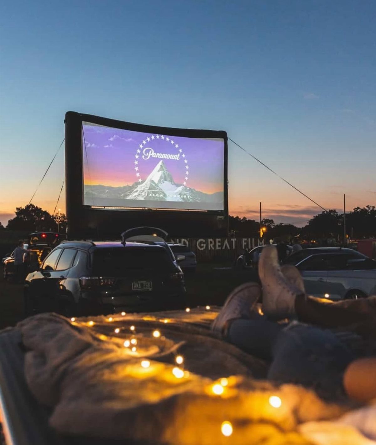Outdoor movie screen at dusk with cars parked in front and cozy blankets.