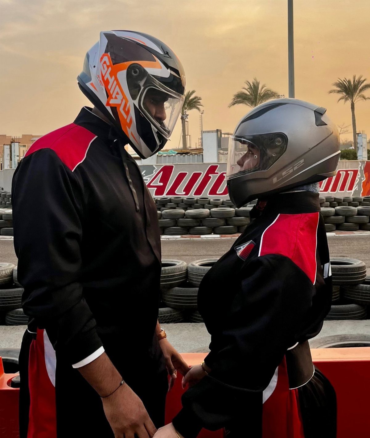 Couple in racing suits and helmets facing each other at a go-kart track.