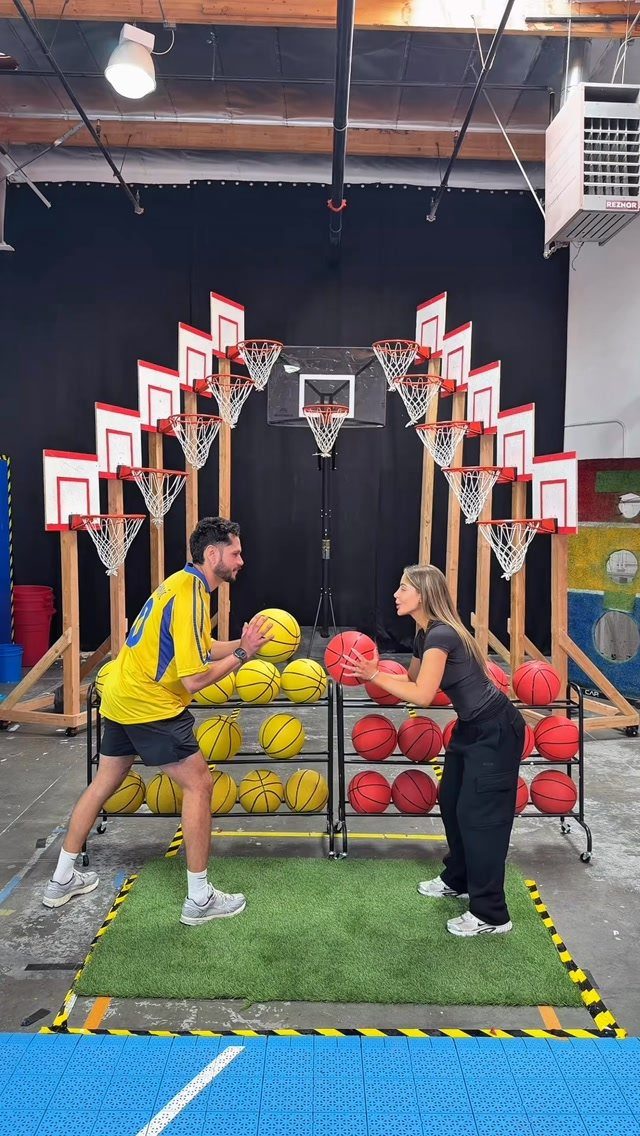 Man and woman holding basketballs in an indoor sports facility with multiple hoops.