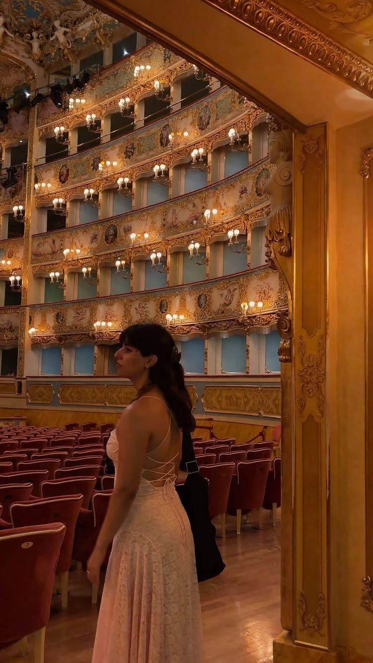 Woman in a formal dress standing inside a lavish, gold-decorated opera house theater.