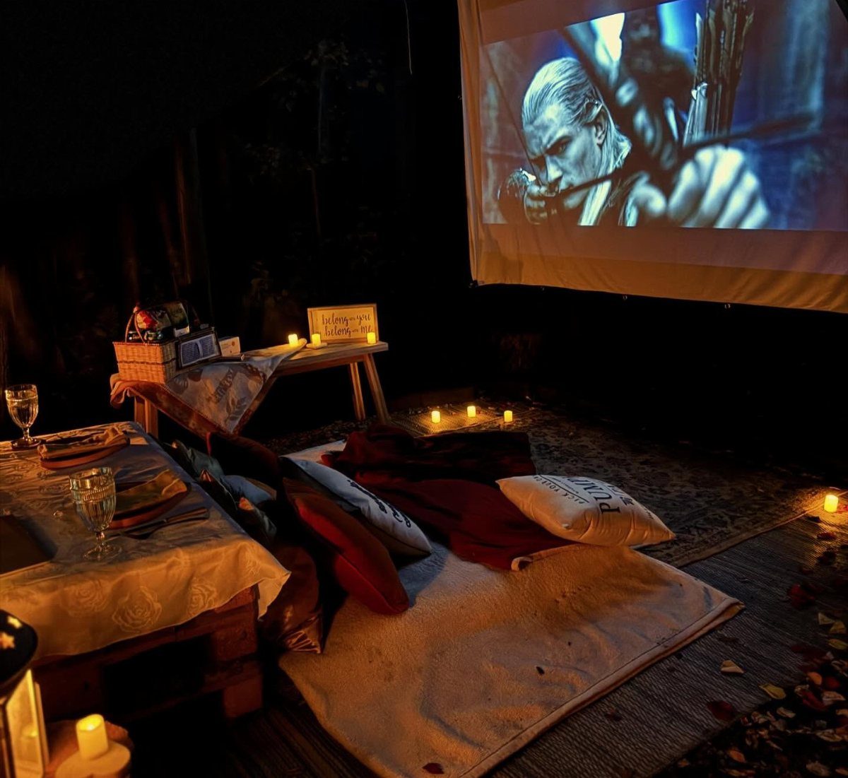 Outdoor movie screen with a cozy mattress, red blankets, and candles at night.