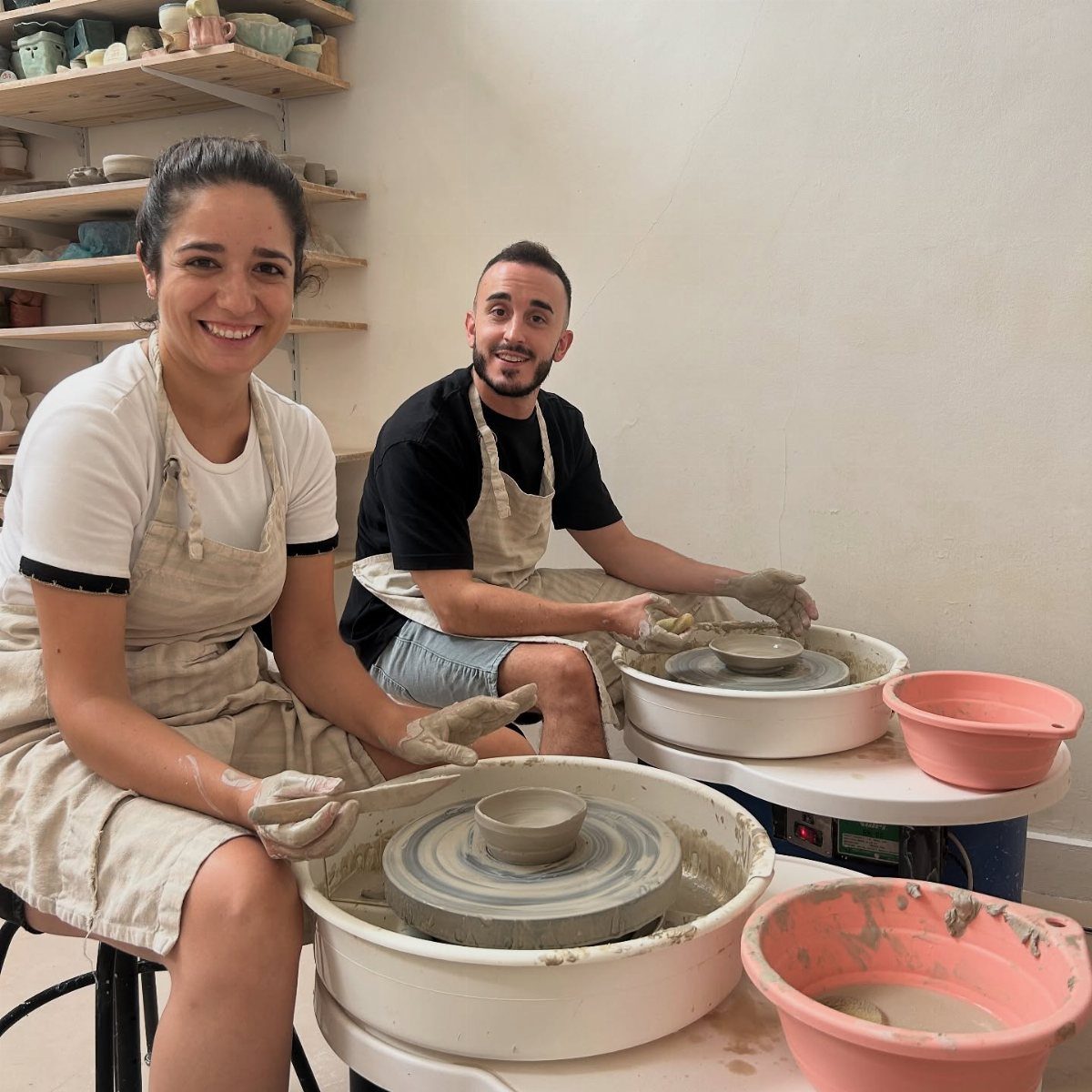 Man and woman in aprons shaping clay bowls on pottery wheels in a studio