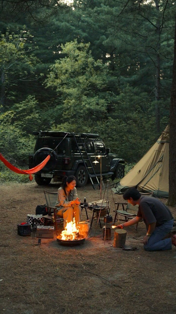 Couple sitting by a campfire next to a tent and off-road vehicle in woods.