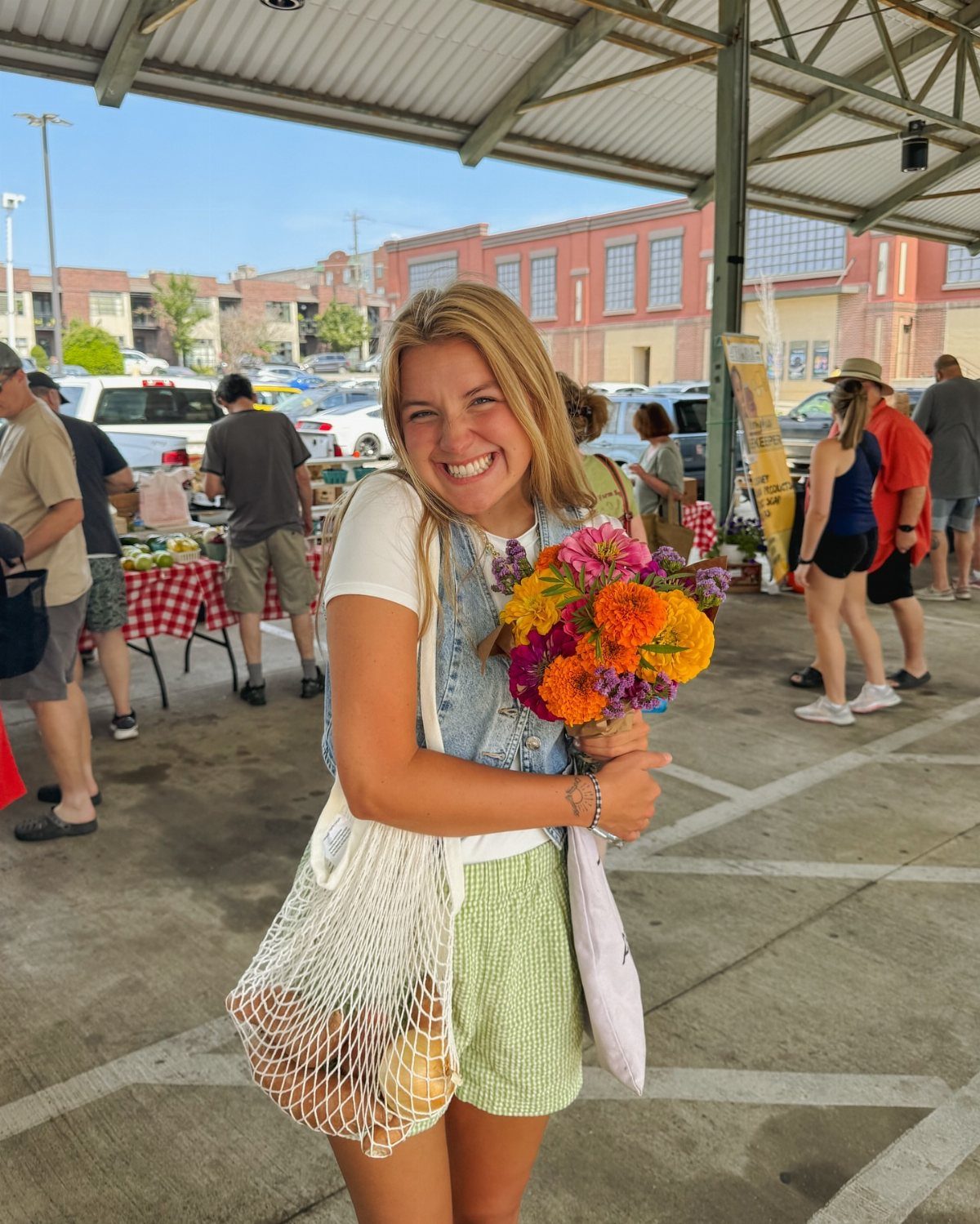 Smiling woman holding a colorful flower bouquet and mesh bag at outdoor market