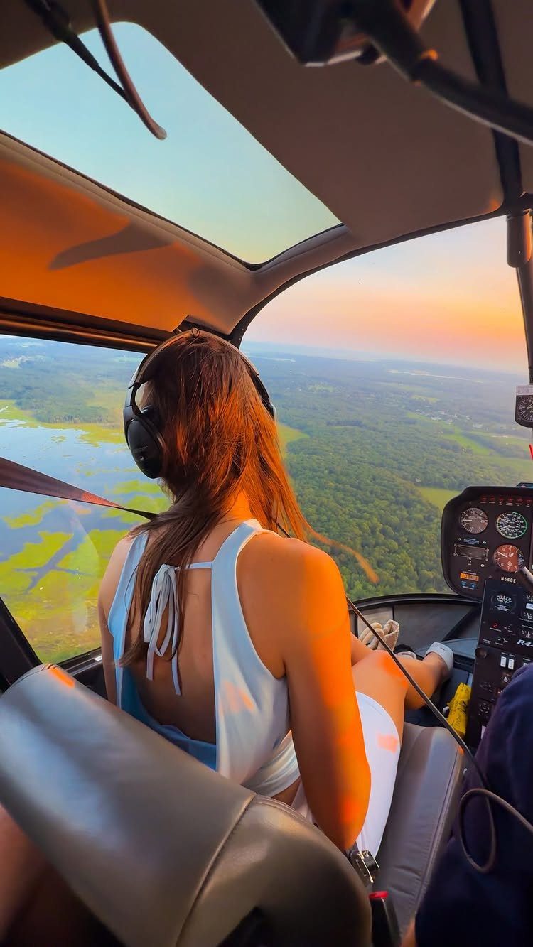 Woman in a white dress wearing headphones inside a helicopter overlooking a sunset landscape.