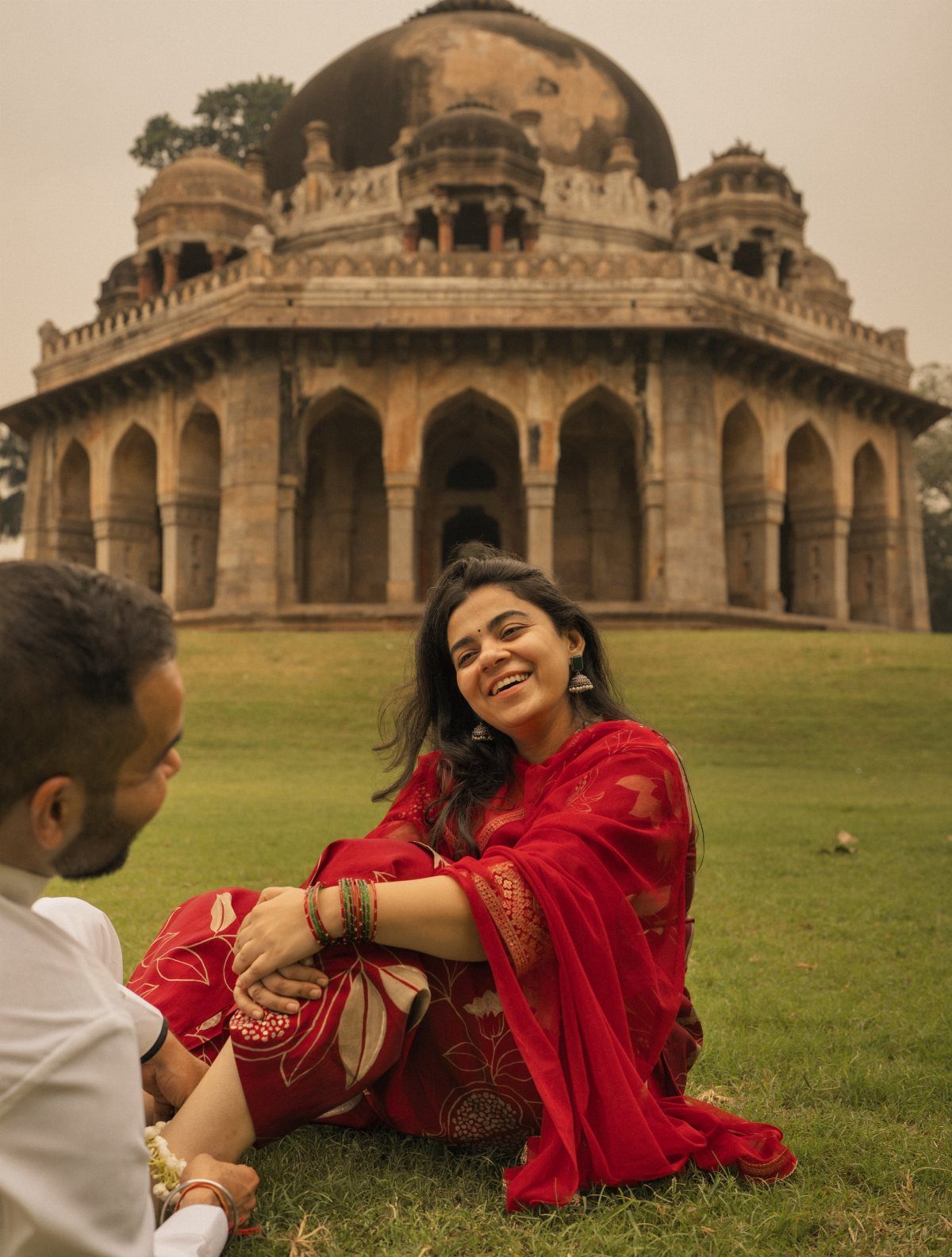 Couple in traditional Indian clothing sitting on grass before a historic domed monument.