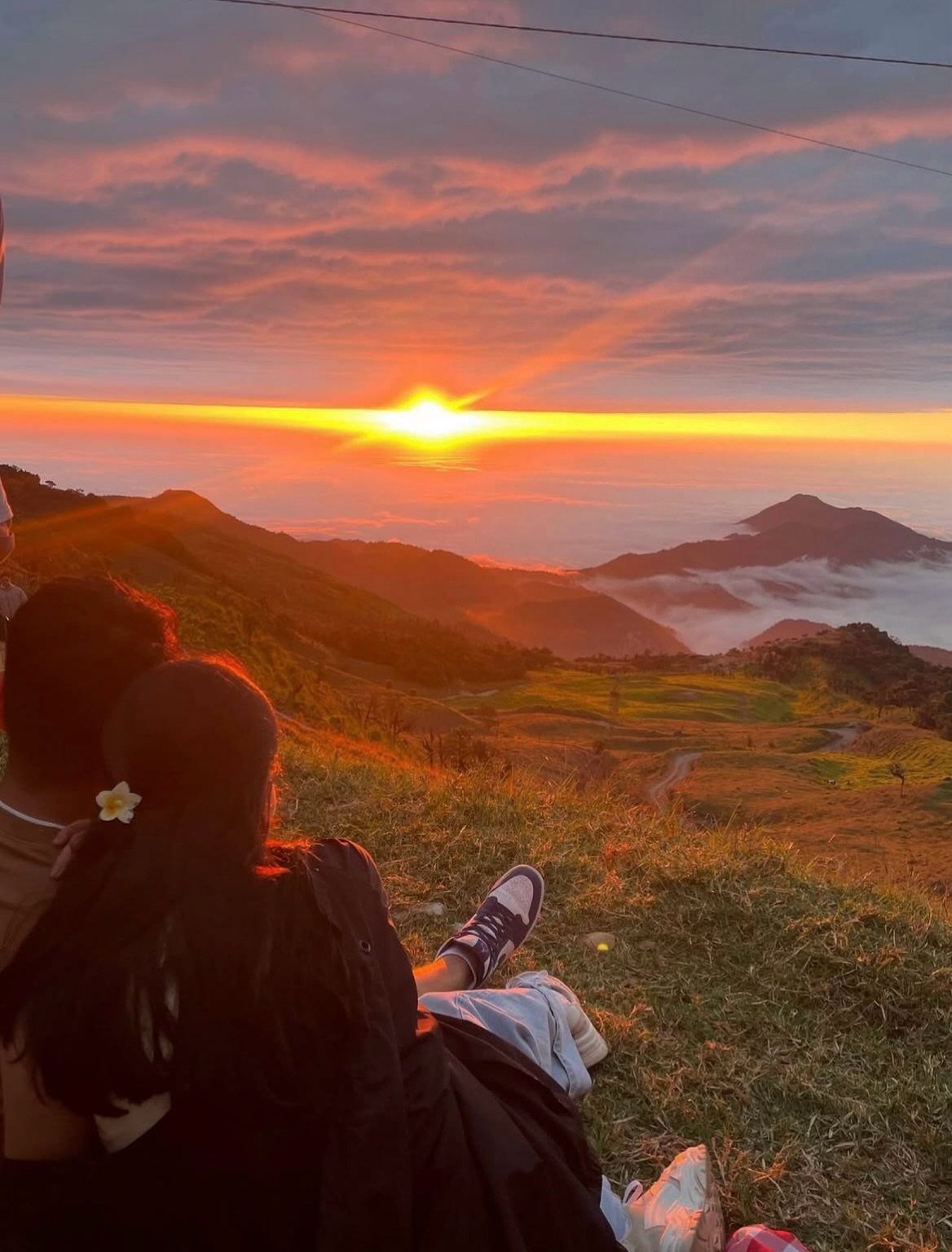 Couple sitting on a grassy hill watching a vibrant orange sunset over mountains.