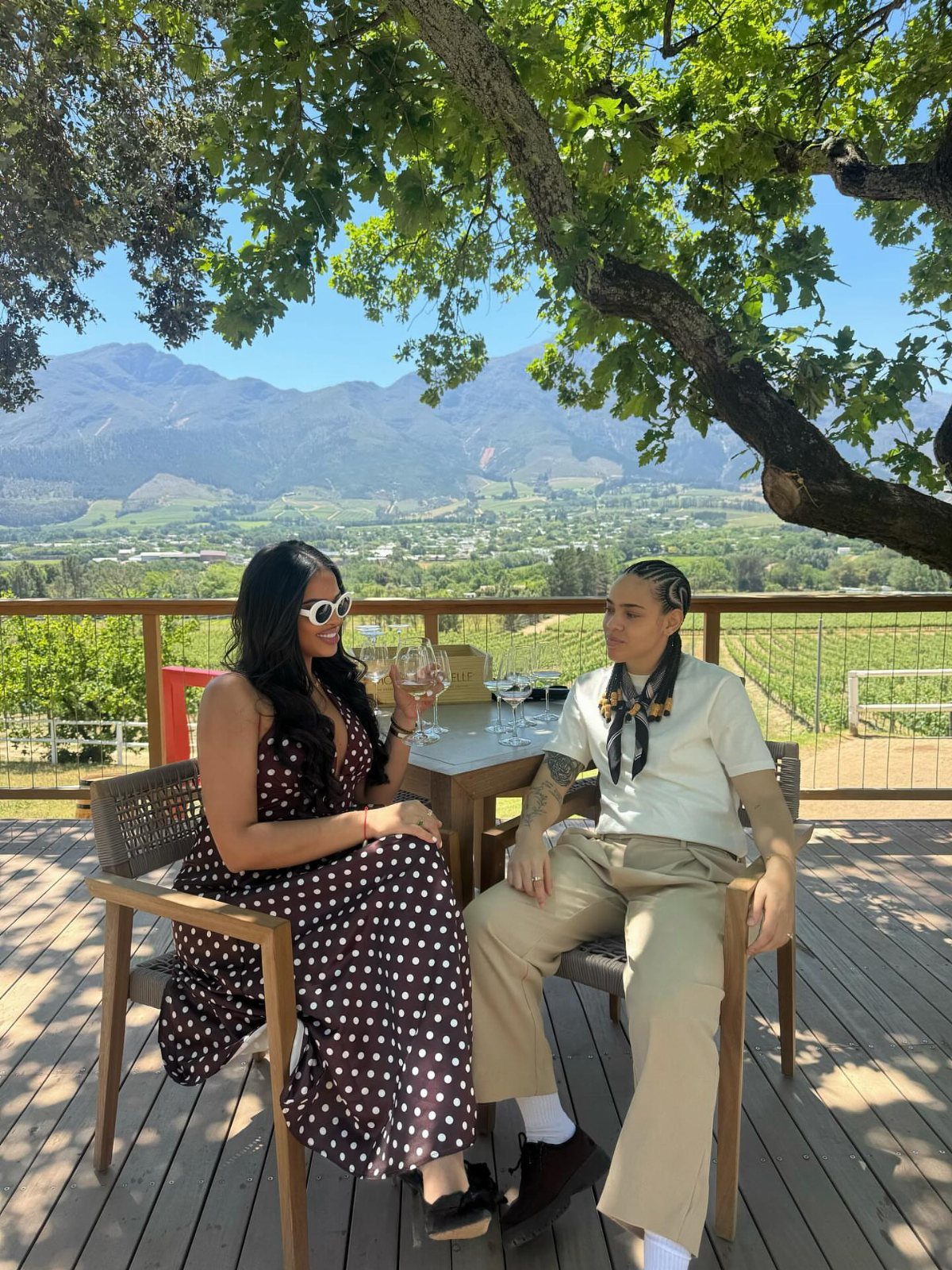 Two people enjoying wine at a table overlooking a scenic vineyard and mountains.