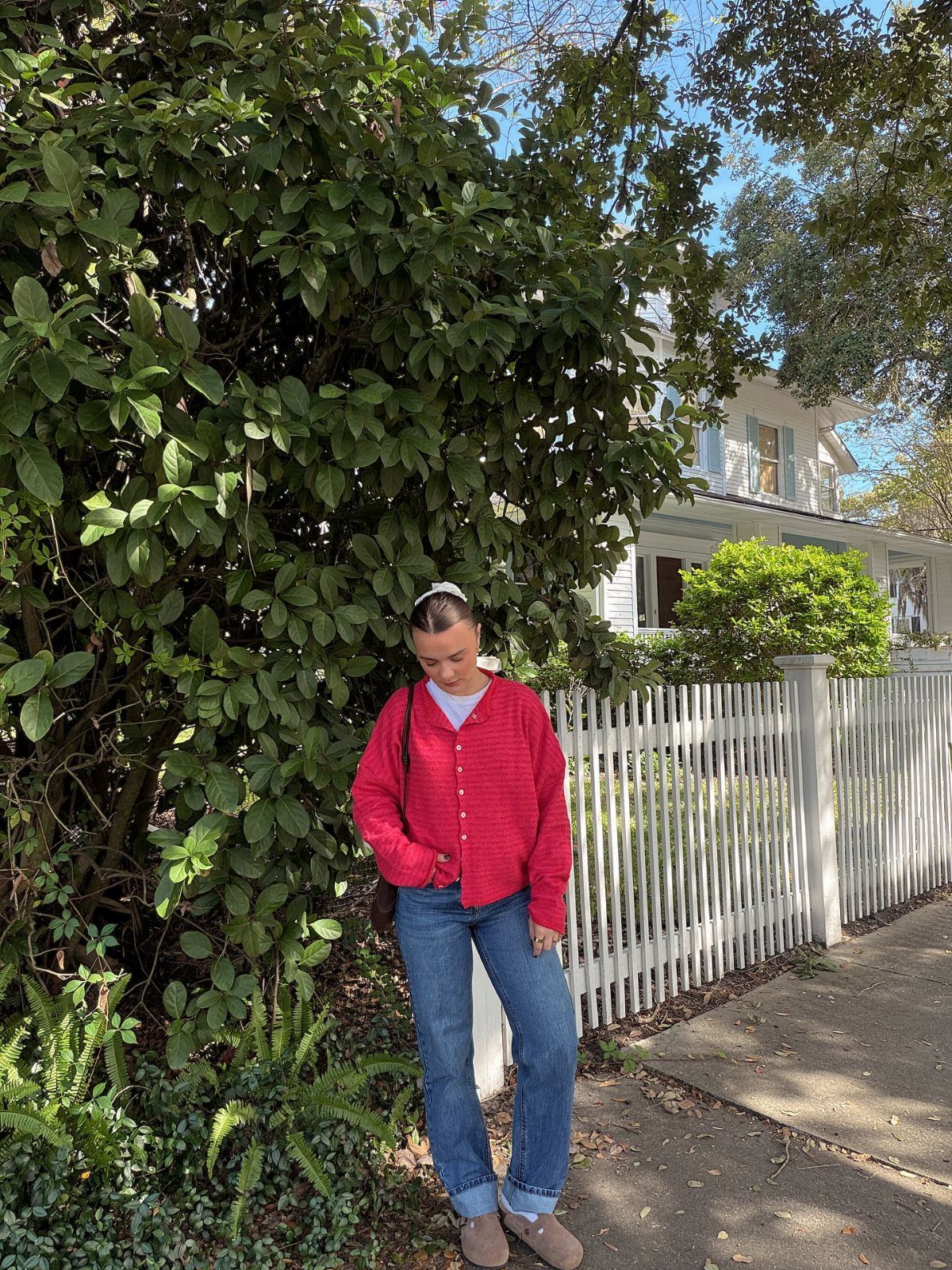 Woman wearing a red cardigan and blue jeans standing by a white fence.