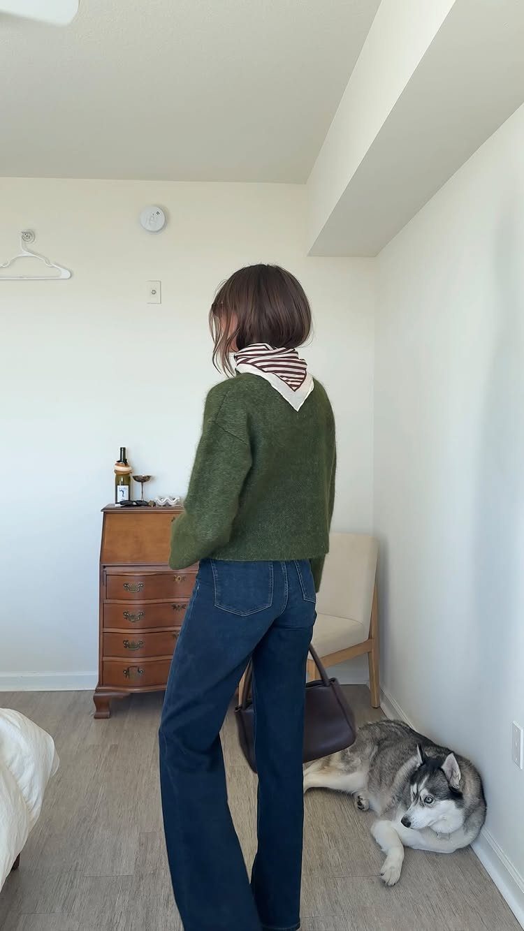 Person wearing olive green sweater and wide-leg jeans standing in a white room.