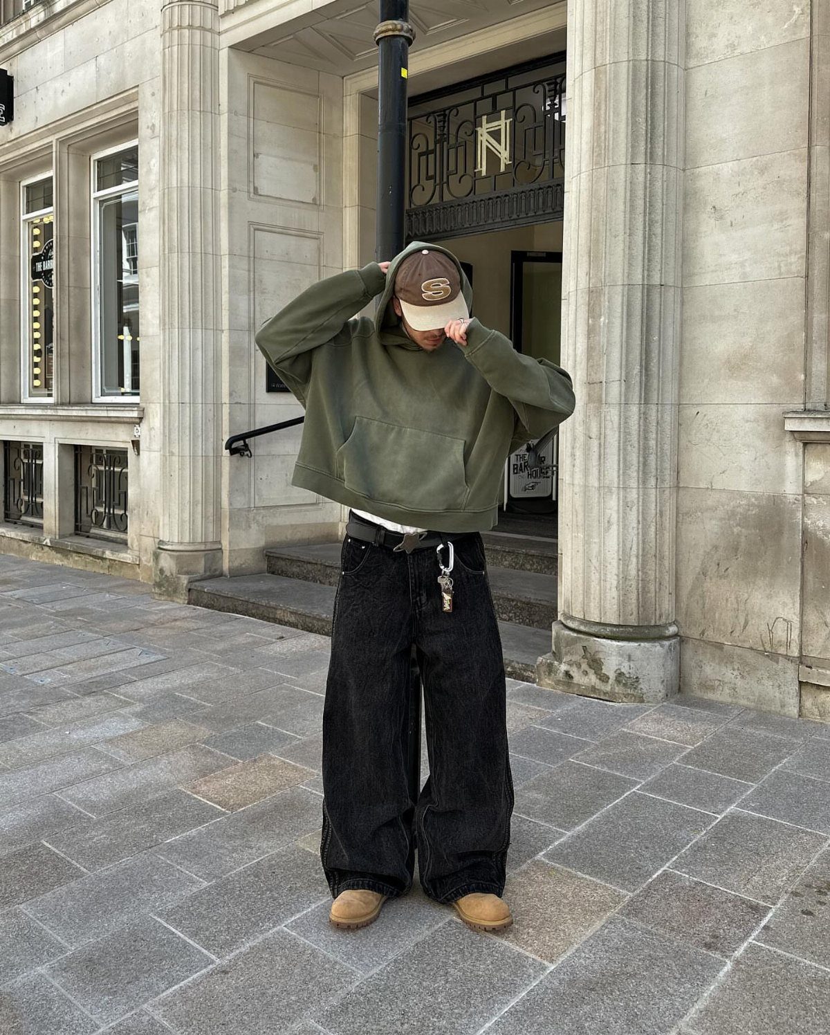 Person wearing an olive green hoodie and wide-leg black jeans on a city street.