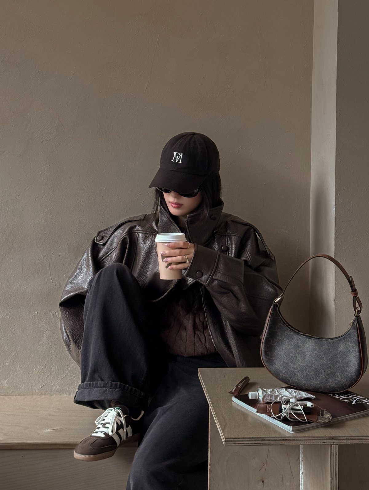 Person in oversized brown leather jacket, black cap, and wide-leg trousers holding coffee.