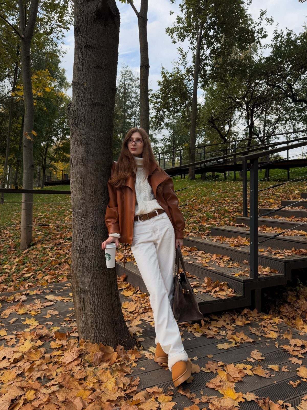 Woman in brown leather jacket and white trousers leaning against a tree in autumn.