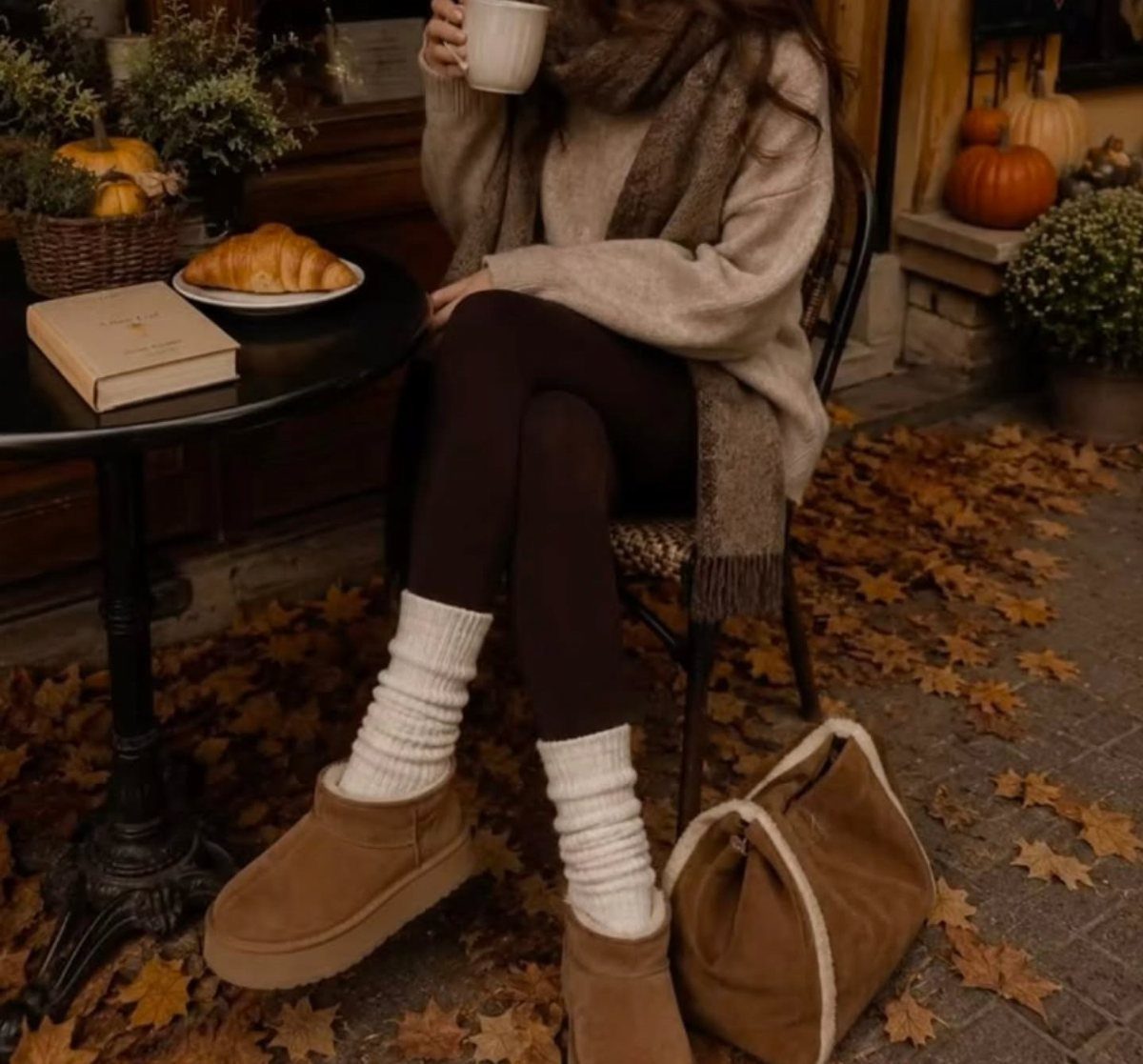 Person in beige sweater and tan boots sitting at cafe with autumn leaves