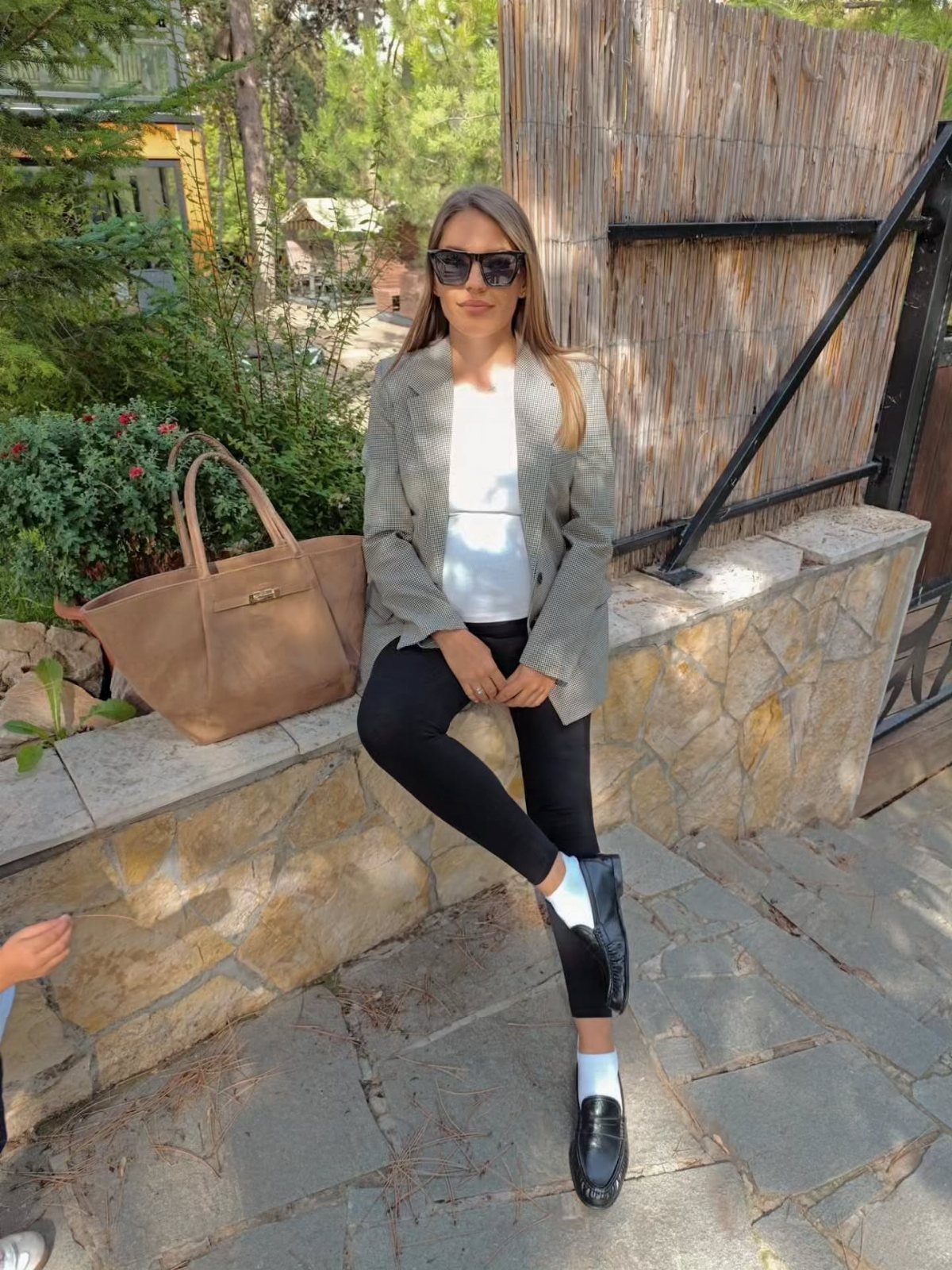 Woman in a grey blazer and black loafers sitting on a stone ledge.