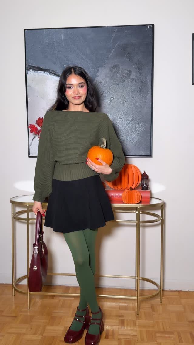 Woman in olive sweater and black skirt holding a pumpkin in a room