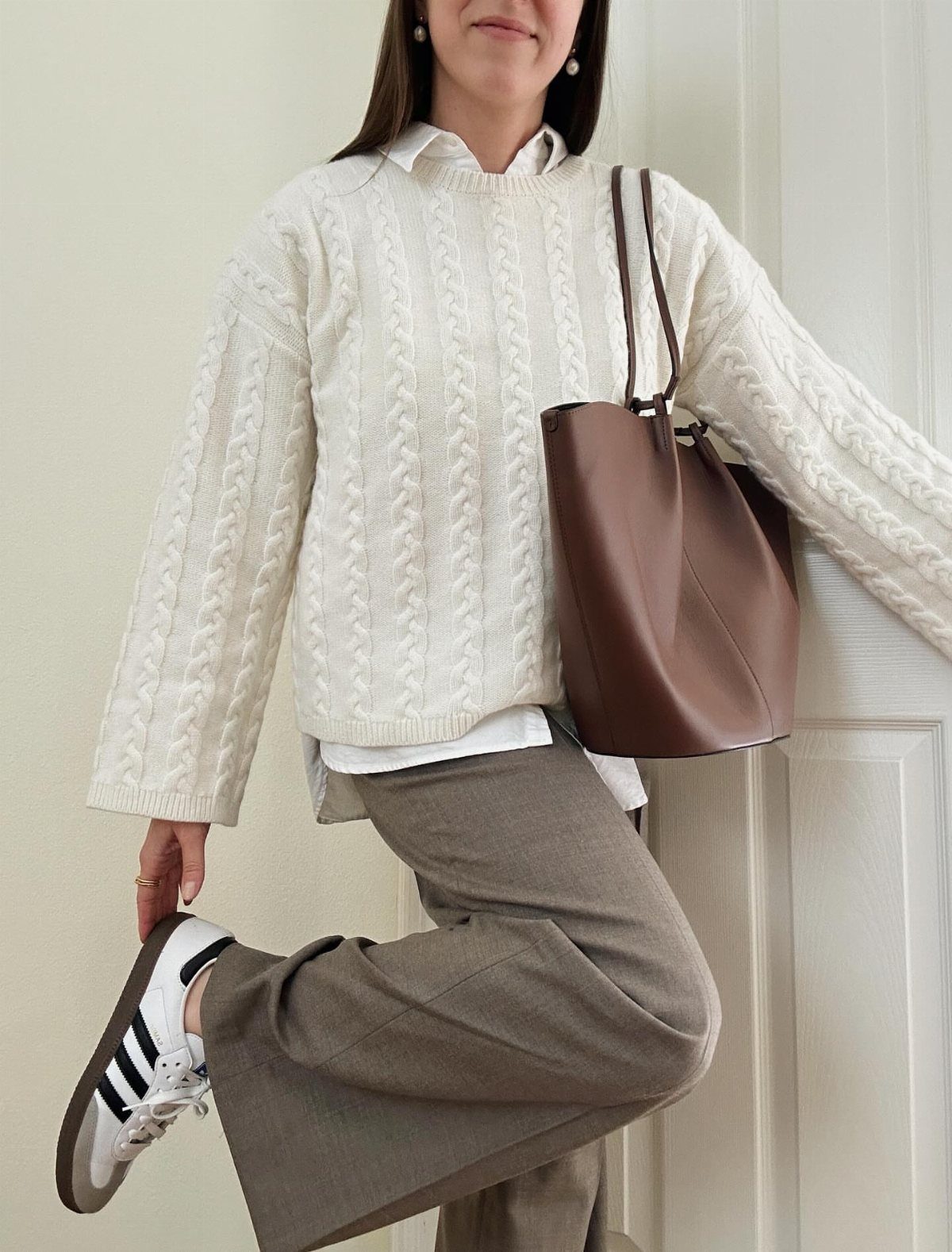 Person in white cable-knit sweater and taupe wide-leg trousers holding a brown tote bag.