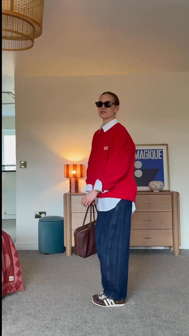 Woman wearing a red sweater over a white shirt and blue trousers indoors.