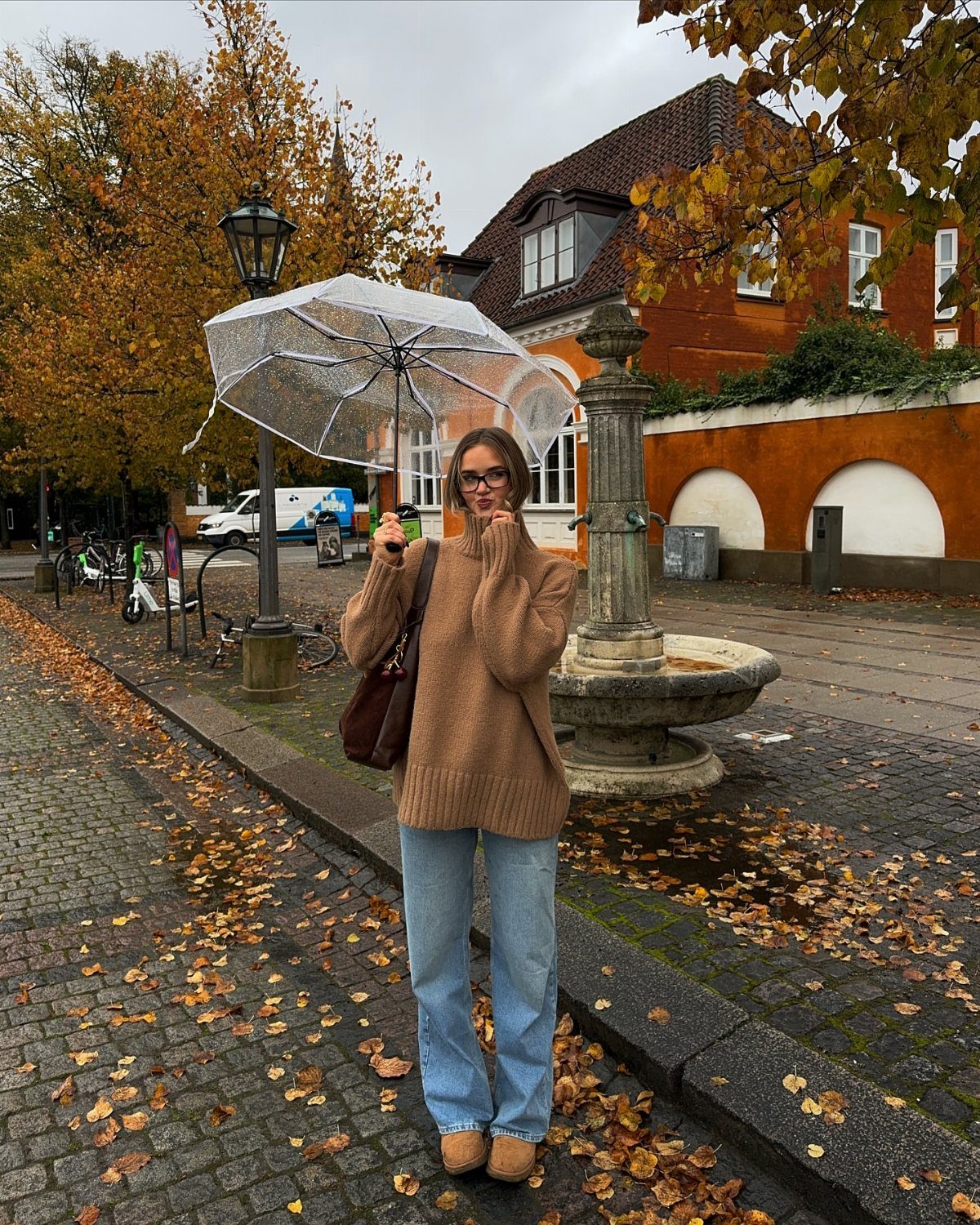 Woman in tan oversized sweater and blue jeans holding a clear umbrella outdoors.