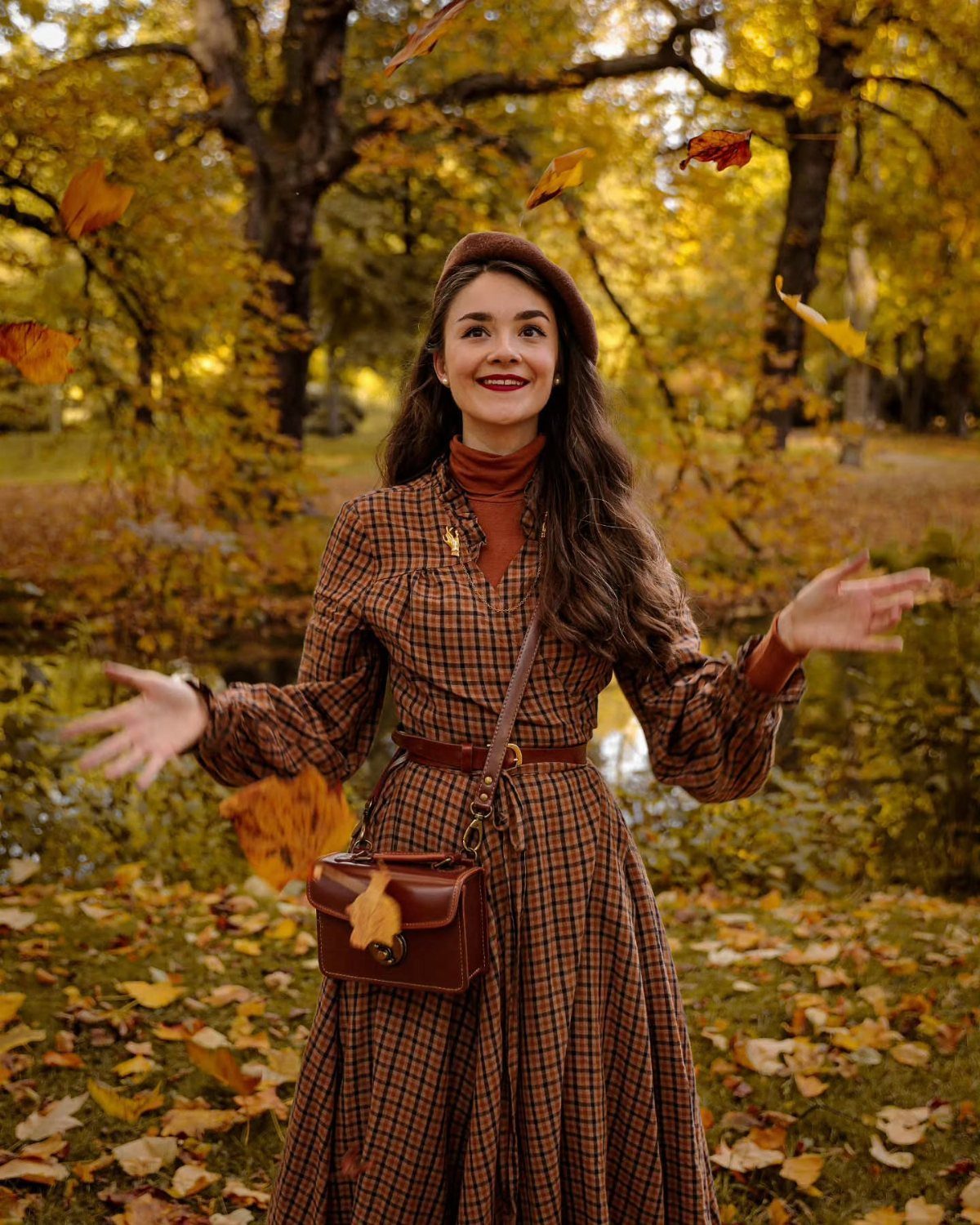 Woman in brown plaid shirtdress and beret standing in autumn park with falling leaves