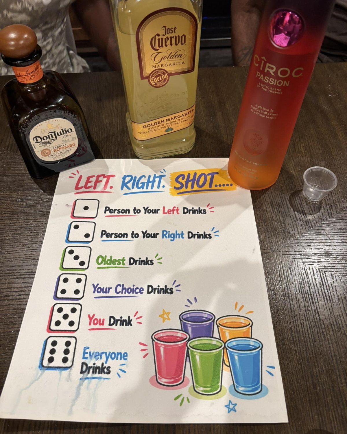 Drinking game rule sheet with liquor bottles and a shot glass on a table