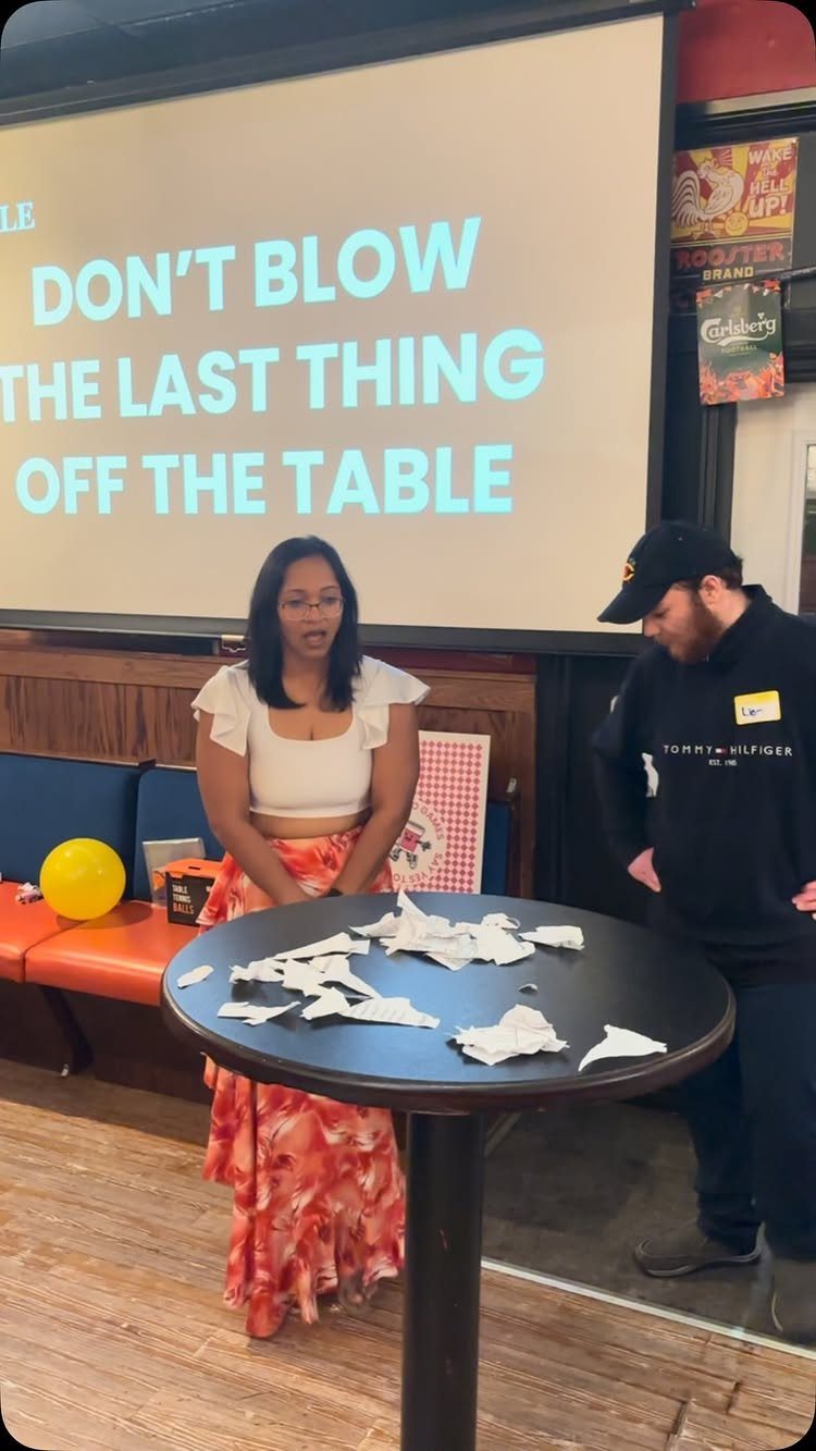 Woman and man standing by a table with paper scraps and a game screen.