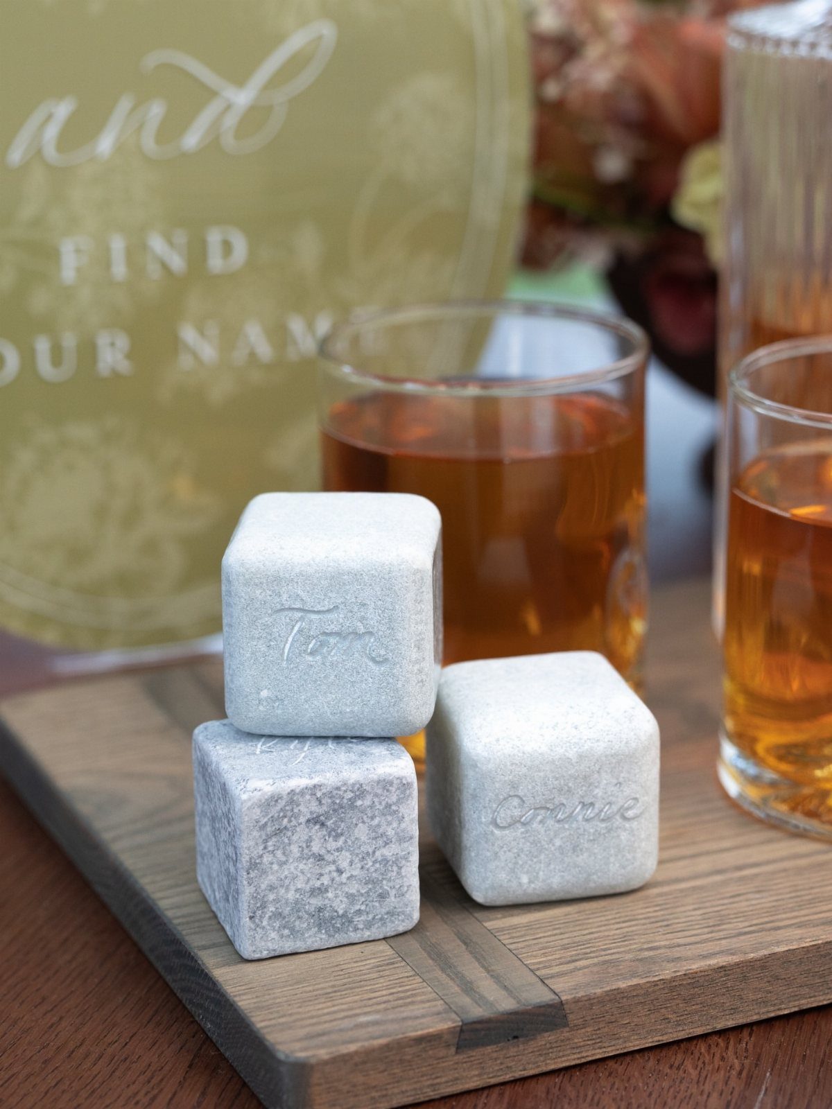 Engraved grey whiskey stones stacked on a wooden board beside glasses of liquor.