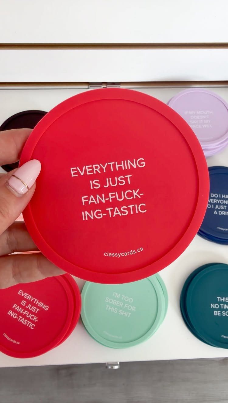 Red circular coaster with humorous white text held by a hand over other coasters.
