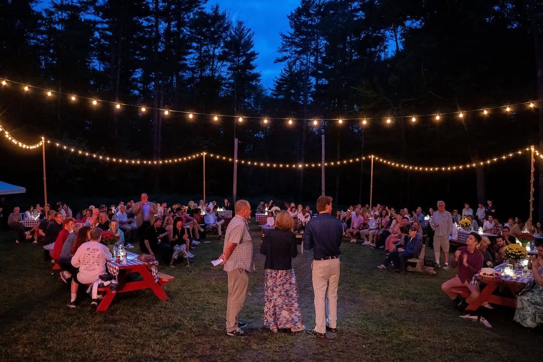 Adults socializing outdoors at dusk under hanging string lights in a wooded area.
