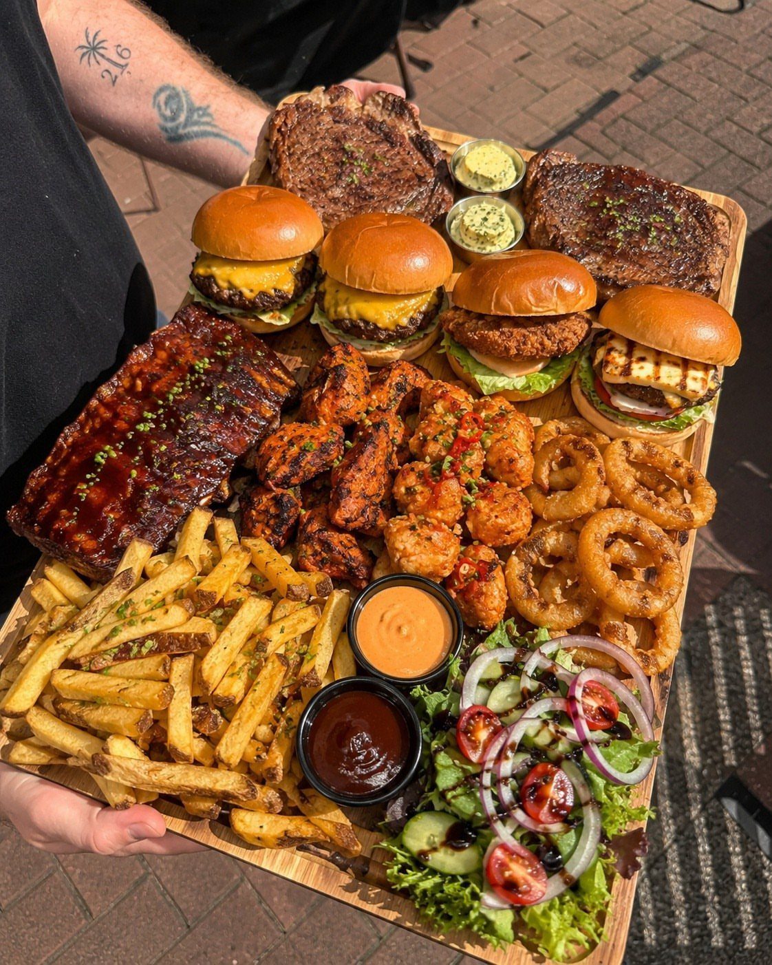 Large wooden platter with sliders, ribs, wings, fries, and steaks held by a person.