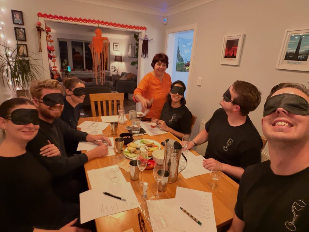Blindfolded adults sitting at a table with wine glasses for a tasting game.
