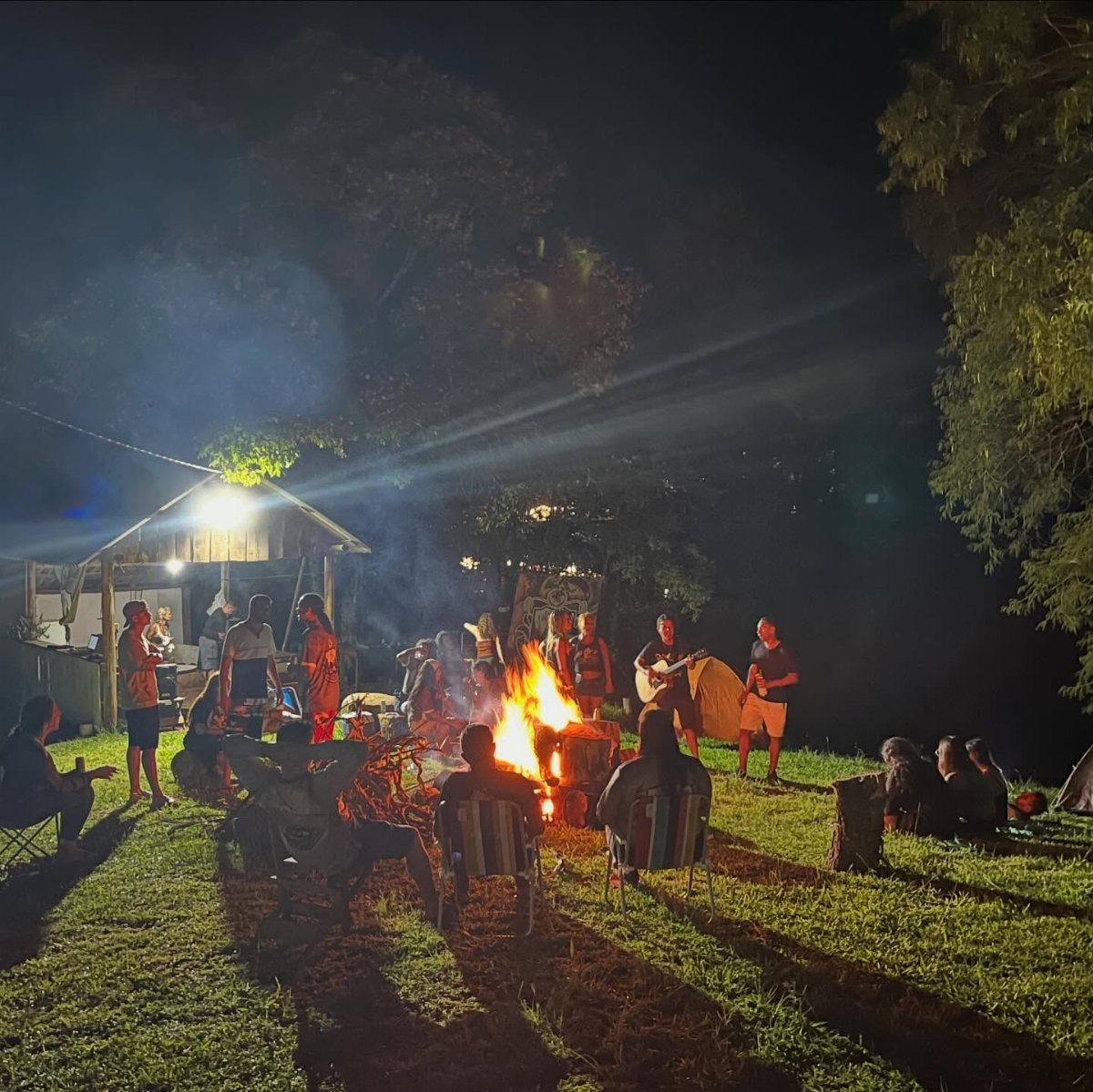 People gathering around a glowing campfire at night with a guitar player.