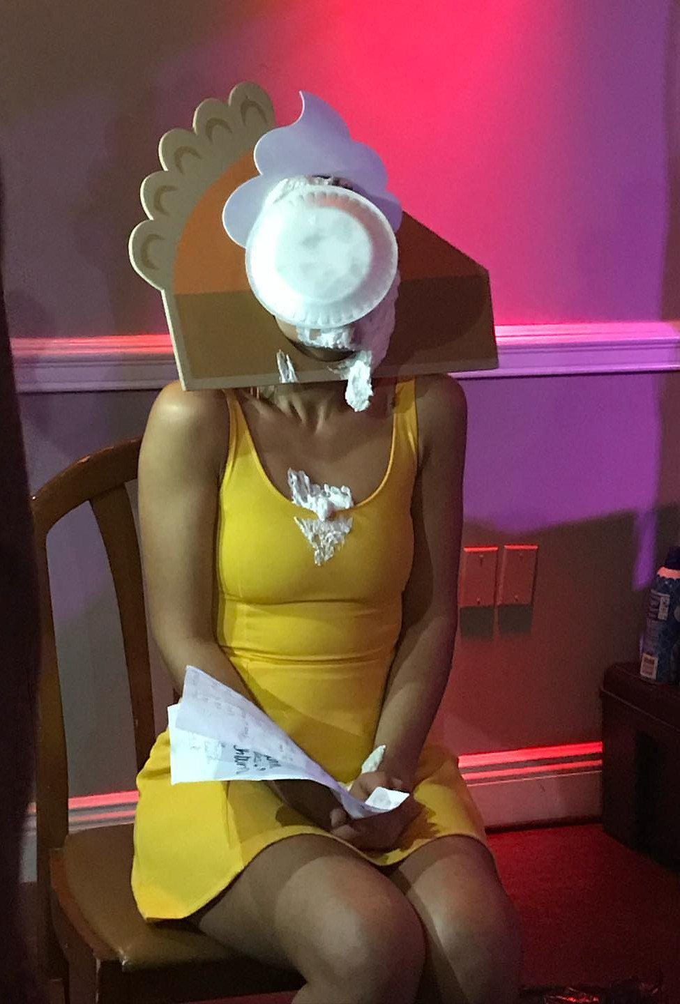 Person in a yellow dress wearing a large cardboard pie slice head costume.
