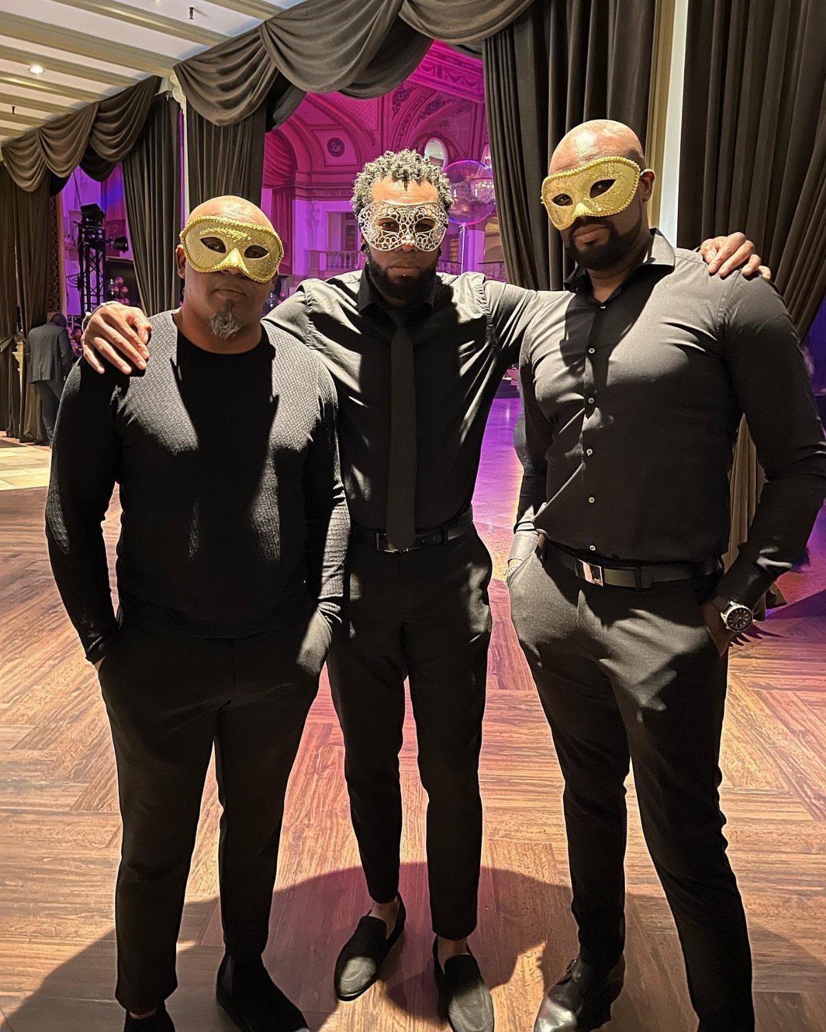 Three men in black outfits wearing gold masquerade masks posing together indoors.