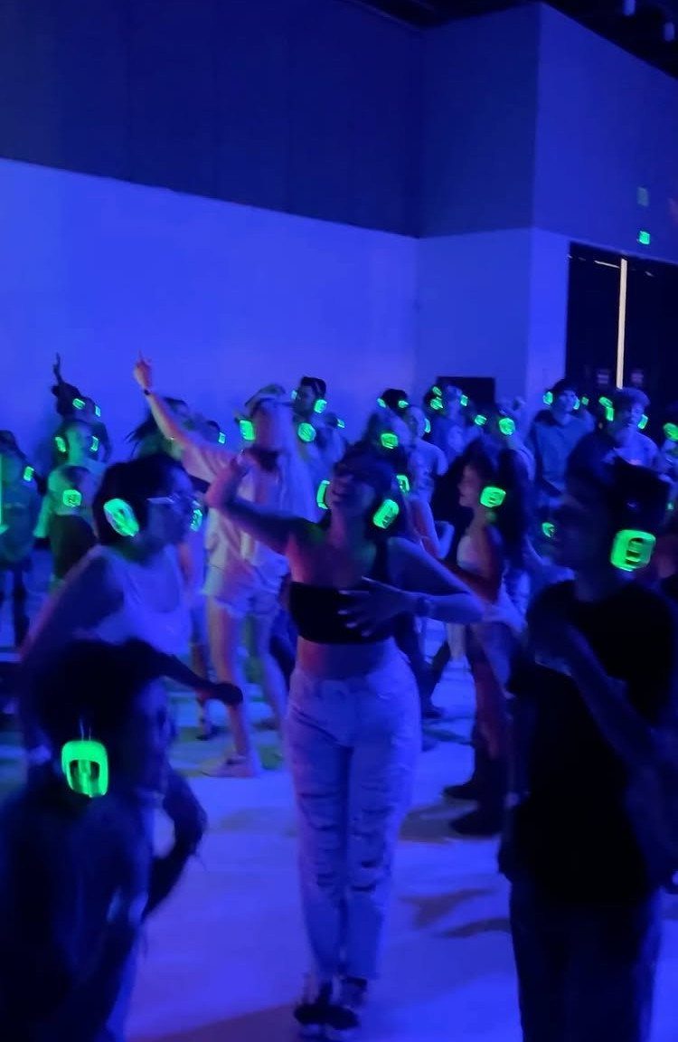 People dancing in a blue-lit room wearing glowing green silent disco headphones.
