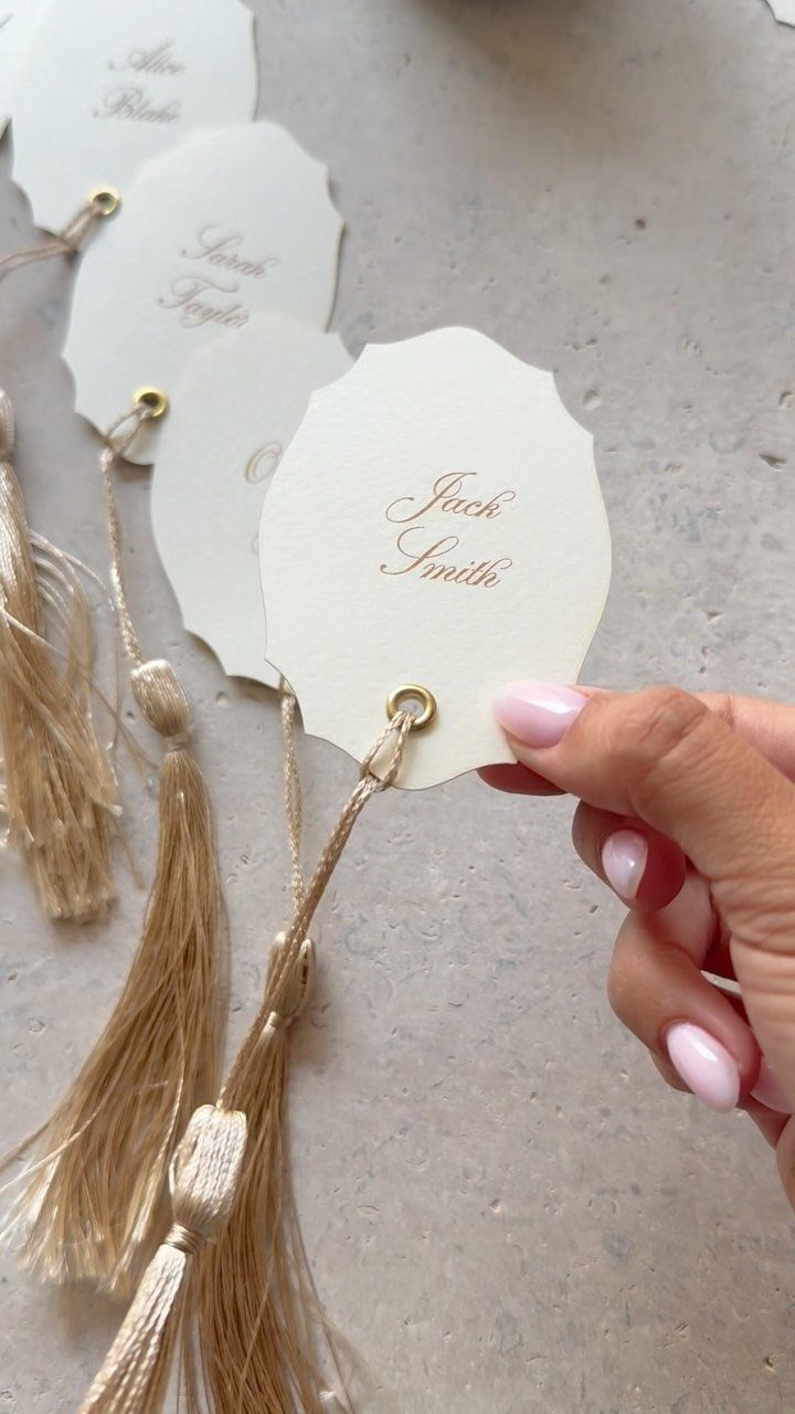 Hand holding a white scalloped name tag with gold cursive text and tassels.