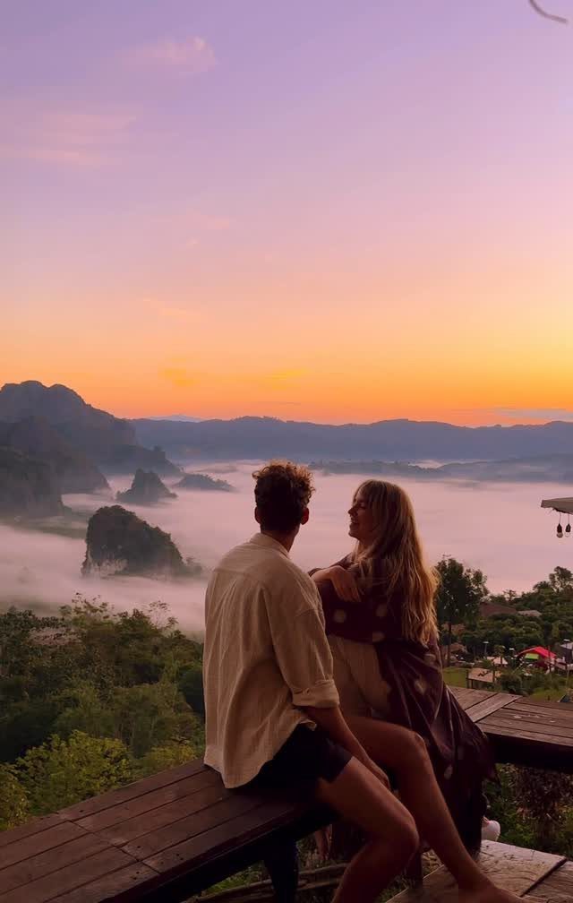 Couple sitting on a wooden deck overlooking misty mountains during a colorful sunset.